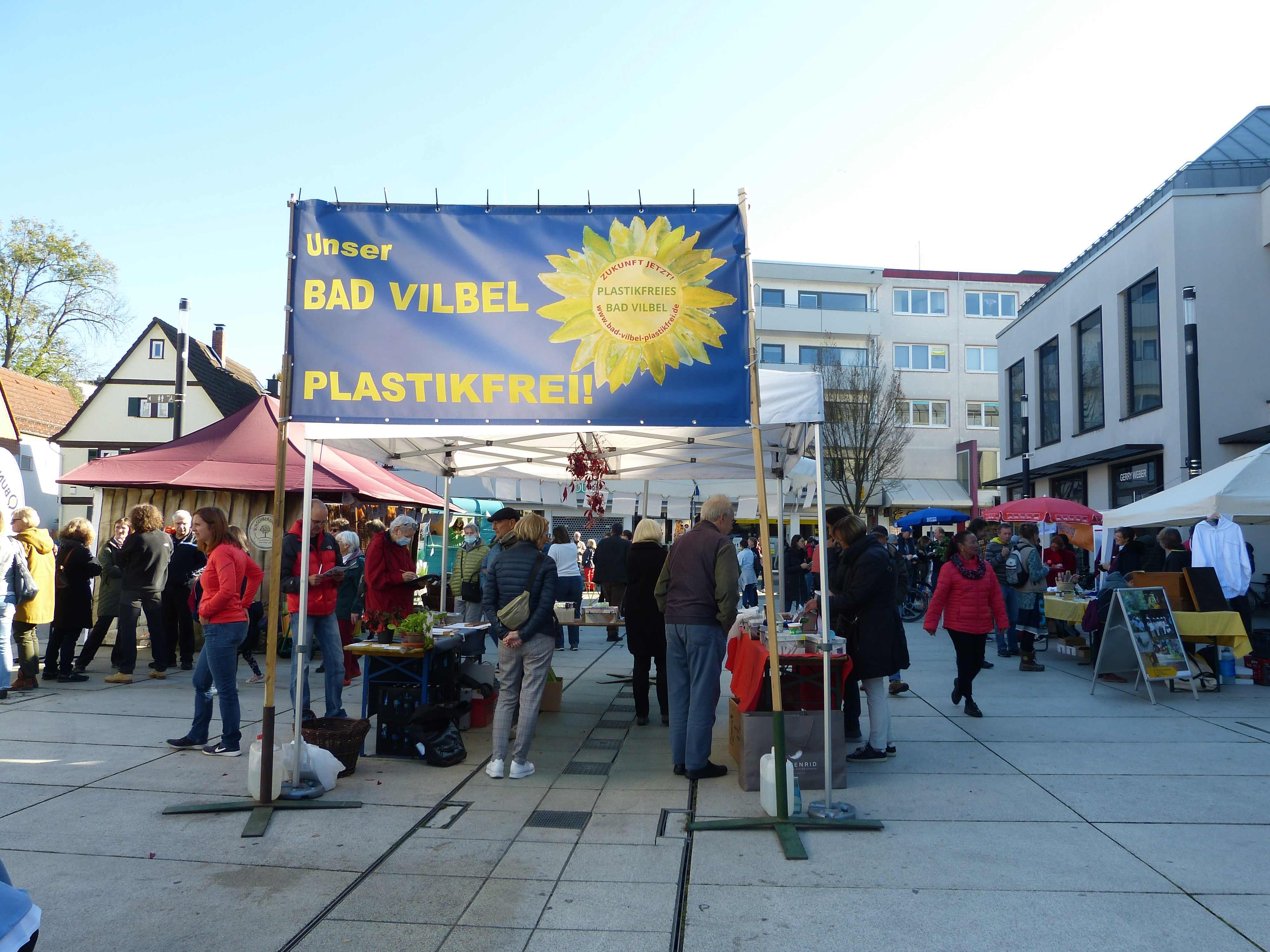 Nachhaltigkeitsmesse Messestand mit blauem Banner 'Unser BAD VILBEL PLASTIKFREI' und mehreren Menschen auf einem Platz zwischen Gebäuden
