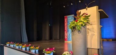 Bühne mit Rednerpult, großem Blumenarrangement und mehreren kleinen Blumentöpfen am Bühnenrand, im Hintergrund ein Banner mit der Aufschrift 'Sportlerehrung Bad Vilbel'