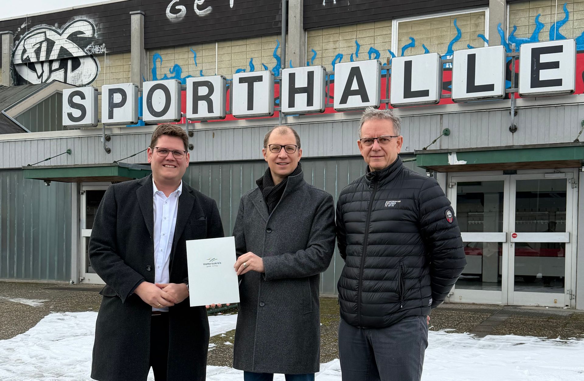 Drei Männer in Winterkleidung stehen vor einem Gebäude mit dem Schriftzug Sporthalle, einer hält eine Mappe