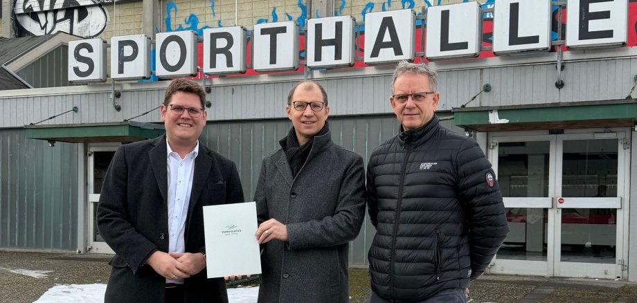 Sporthallenneubau Drei Männer in Winterkleidung stehen vor einem Gebäude mit dem Schriftzug Sporthalle, einer hält eine Mappe