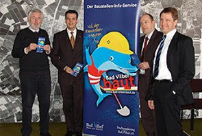 Vier Männer in Anzügen stehen vor einer Wand mit Stadtplan und einem Roll-Up mit einer blauen Ente in Bauarbeiterkleidung darauf