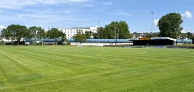 Weitläufiges Sportfeld mit gepflegtem Rasen, überdachter Tribüne und Bäumen im Hintergrund unter blauem Himmel