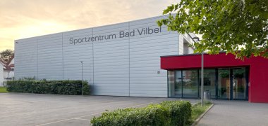 Außenansicht des Sportzentrums Bad Vilbel mit grauer Fassade, rotem Eingangsbereich und Parkplatz im Vordergrund