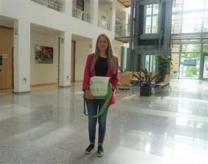 Frau steht in einem hellen, modernen Foyer und hält eine grüne Tasche mit Aufschrift 'Familienbüro'