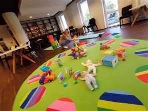 Kind spielt auf grünem Teppich mit bunten Kreisen in einem Raum mit Bücherregal und großen Fenstern
