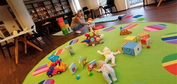 Familienbüro Kind spielt auf grünem Teppich mit bunten Kreisen in einem Raum mit Bücherregal und großen Fenstern