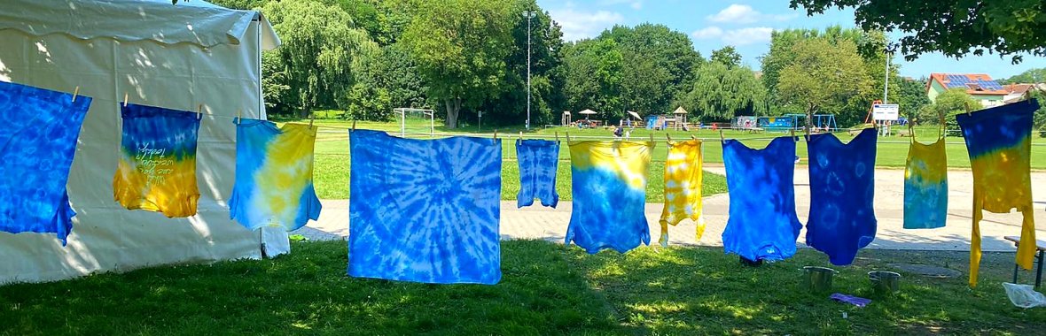 Mehrere blaue und gelbe Batik-Tücher hängen an einer Wäscheleine im Freien vor einer Wiese mit Bäumen und Spielplatzgeräten