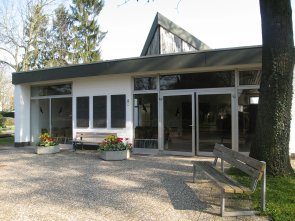 Trauerhalle mit großen Glasfenstern, davor zwei Blumenkübel mit roten und gelben Blumen und eine Holzbank neben einem Baum