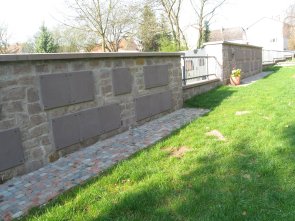 Steinerne Mauer (Urnenwand) mit mehreren rechteckigen Metallplatten und gepflastertem Weg daneben auf einer Wiese in einem Friedhof