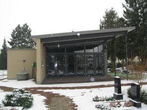 Trauerhalle mit Glasfront und Flachdach auf verschneitem Friedhof mit Grabsteinen und Büschen