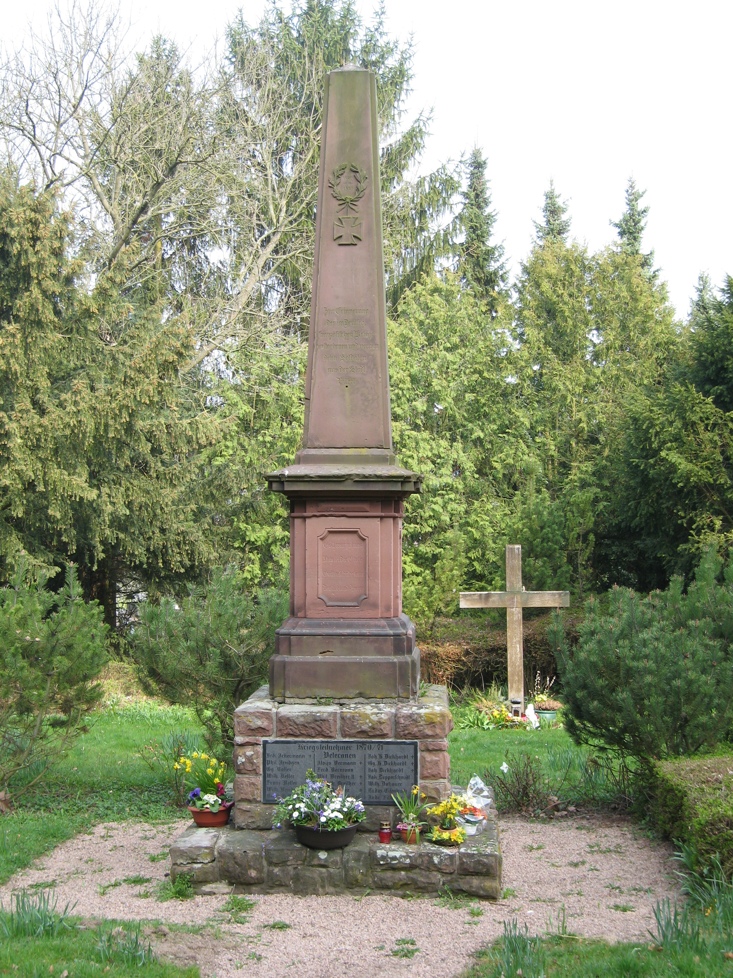 Obelisk auf einem Steinsockel mit Gedenktafeln und Blumen auf einem Friedhof, im Hintergrund ein Holzkreuz und Nadelbäume