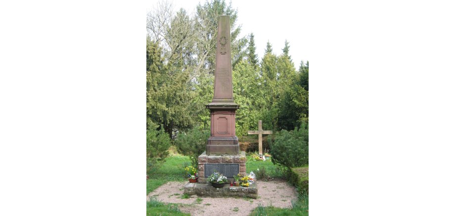 Obelisk auf einem Steinsockel mit Gedenktafeln und Blumen auf einem Friedhof, im Hintergrund ein Holzkreuz und Nadelbäume