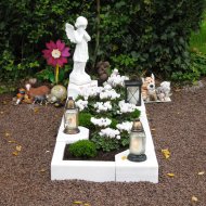 Grab mit weißem Engel als Statue, weißen Blumen, zwei Laternen und mehreren kleinen Tierfiguren vor einem Hintergrund aus Efeu