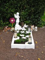 Grab mit weißem Engel als Statue, weißen Blumen, zwei Laternen und mehreren kleinen Tierfiguren vor einem Hintergrund aus Efeu