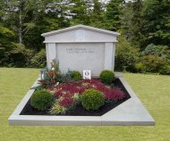 Grabstätte mit rechteckigem, steinernem Grabstein und bepflanztem Grabfeld mit Buchsbaumkugeln und roten sowie grünen Pflanzen auf einem Friedhof