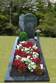 Grab mit schwarzem Grabstein, darauf eine Blume als Relief, davor ein Beet mit roten und weißen Blumen sowie ein Strauß in einer Vase
