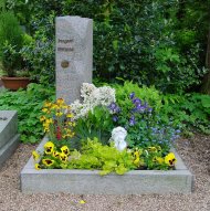 Grabstein umgeben von bunten Blumen und einer kleinen Engelsfigur auf einem gepflegten Friedhof