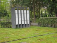 Urnenwand mit mehreren vertikalen Nischen auf einer Rasenfläche auf einem Friedhof mit Bäumen und Hecken im Hintergrund