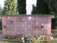 Urnenwand auf einem Friedhof mit mehreren Gedenktafeln, Blumen und Pflanzen davor, umgeben von Bäumen