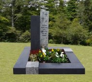 Moderner Grabstein auf einem rechteckigen Grab mit Blumen und Kerze vor Waldkulisse auf einem Friedhof