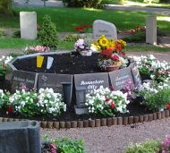 Urnengrab mit mehreren Grabplatten in einem kreisförmigen Arrangement, umgeben von weißen und roten Blumen auf einem Friedhof