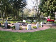 Urnengrabfeld mit mehreren unterschiedlich geformten Grabsteinen in einem kreisförmigen Arrangement auf einem Friedhof, umgeben von Rasen und Bäumen