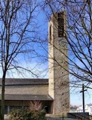 Kirchturm aus hellem Stein mit rechteckigen Fenstern neben einem Gebäude und kahlen Bäumen im Vordergrund