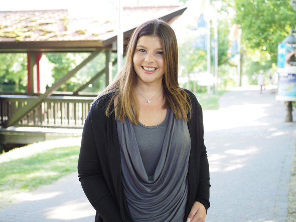 Frau mit langen Haaren, grauem Shirt mit Wasserfallausschnitt und schwarzer Strickjacke steht auf einem Gehweg im Freien