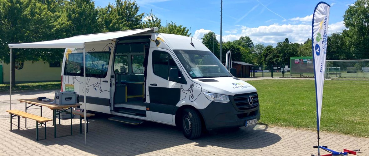 Weißer Mercedes-Sprinter mit ausgefahrener Markise, daneben ein Tisch mit zwei Bänken auf gepflastertem Platz einer Sportanlage. Rechts ein Fahnenbanner und zwei bunte Hula Hoop Reifen auf dem Boden