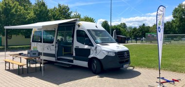 KinderMobil Weißer Mercedes-Sprinter mit ausgefahrener Markise, daneben ein Tisch mit zwei Bänken auf gepflastertem Platz einer Sportanlage. Rechts ein Fahnenbanner und zwei bunte Hula Hoop Reifen auf dem Boden