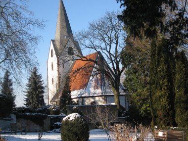Kirche mit spitzem Turm hinter kahlen Bäumen und verschneitem Friedhof mit Grabsteinen und Bänken