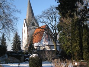 Kirche mit spitzem Turm hinter kahlen Bäumen und verschneitem Friedhof mit Grabsteinen und Bänken