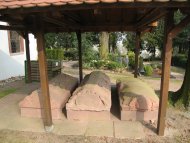Drei steinerne Sarkophage unter einem Holzdach auf einem Friedhof mit Bäumen und Gräbern im Hintergrund