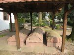 Drei steinerne Sarkophage unter einem Holzdach auf einem Friedhof mit Bäumen und Gräbern im Hintergrund