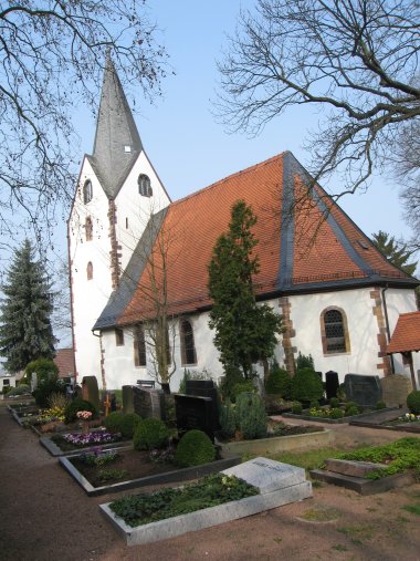 Kirche mit spitzem Turm und rotem Dach umgeben von Grabsteinen und Bäumen auf einem Friedhof