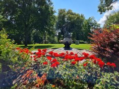Brunnen mit einer Skulptur in einem Park, umgeben von roten Blumen und Bäumen im Hintergrund