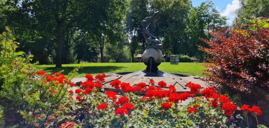 Brunnen mit einer Skulptur in einem Park, umgeben von roten Blumen und Bäumen im Hintergrund
