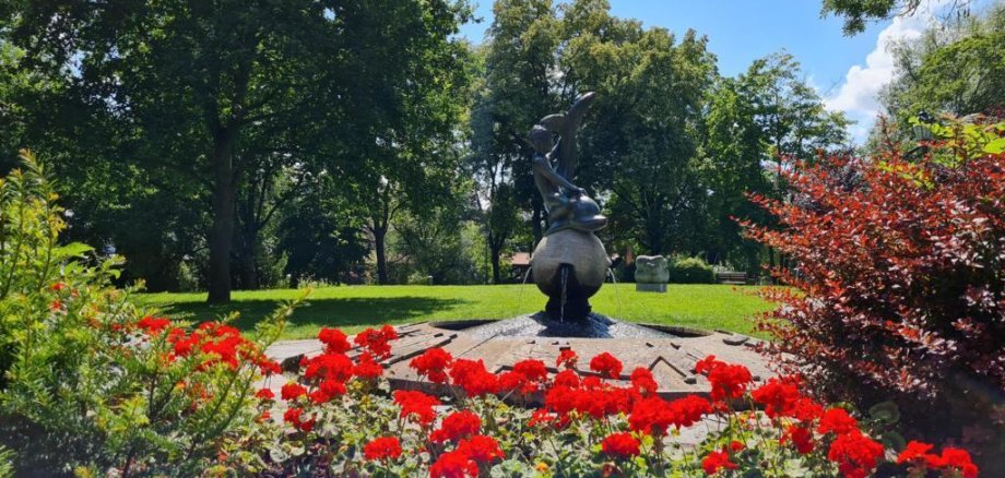 Brunnen Brunnen mit einer Skulptur in einem Park, umgeben von roten Blumen und Bäumen im Hintergrund