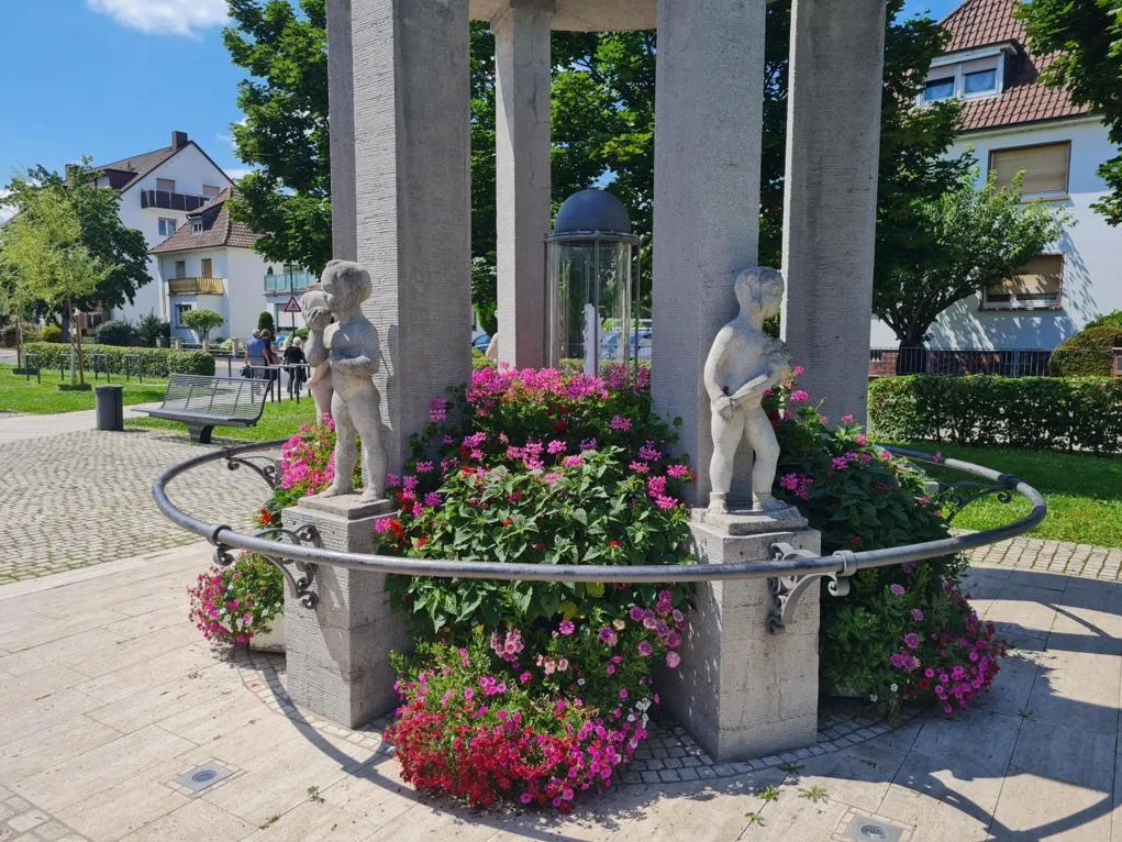 Brunnen mit zwei steinernen Kinderskulpturen, umgeben von Blumen und einem Metallring, im Hintergrund Wohnhäuser und Bänke