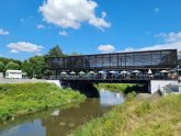 Modernes Gebäude mit Glasfassade und Sonnenschirmen auf einer Brücke über einem Fluss unter blauem Himmel