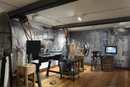 Museumsausstellung mit historischen Maschinen und Geräten auf Holzboden vor einer Wand mit großflächigem schwarz-weiß-Fotodruck