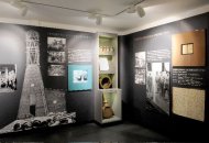 Museumsausstellung mit historischen Fotos, Texttafeln und einem Regal mit alten Gegenständen vor schwarzem Hintergrund