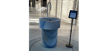 Brunnen Vilco Runder blauer Marmorbrunnen mit gebogenem Metallrohr und Infotafel auf Ständer in hellem Innenraum mit Treppe und Steinwand