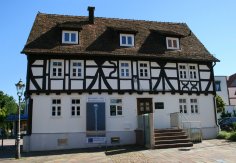 Fachwerkhaus mit weißer Fassade und dunklen Holzbalken, einem Eingang und mehreren Fenstern, davor Pflasterplatz und Laterne