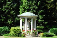 Runder Brunnentempel mit Säulen und einer Statue in einem Park umgeben von Büschen und Bäumen
