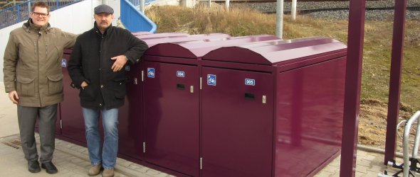 Zwei Männer stehen neben einer Reihe von Fahrradboxen mit geschwungenen Dächern und blauen Rollstuhlsymbolen