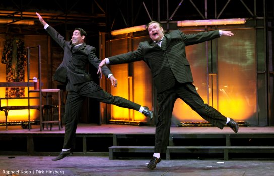 Zwei Männer in Anzügen tanzen synchron auf einer Bühne mit orange beleuchtetem Hintergrund