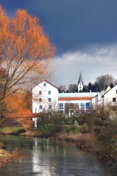 Fluss mit Uferbäumen und weißem Gebäude im Hintergrund, darüber ein Kirchturm unter bewölktem Himmel