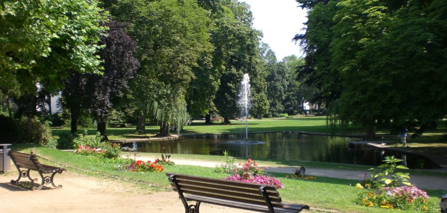 Kurpark Parkbank vor einem Teich mit Springbrunnen umgeben von Bäumen und Blumen im Kurpark