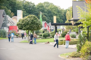 Menschen gehen auf einem breiten Weg zwischen modernen Einfamilienhäusern mit Gärten und Bäumen in einer Musterhaus-Ausstellung